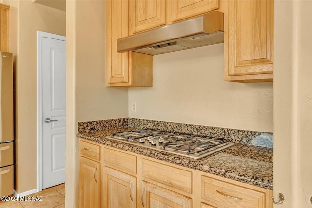 kitchen featuring light tile patterned floors, dark stone countertops, stainless steel gas cooktop, exhaust hood, and light brown cabinetry