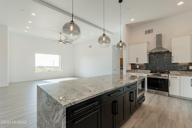 kitchen featuring light wood-type flooring, appliances with stainless steel finishes, white cabinetry, wall chimney range hood, and a kitchen island