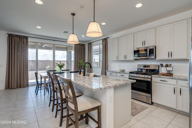 kitchen featuring a kitchen island with sink, appliances with stainless steel finishes, hanging light fixtures, light stone countertops, and white cabinetry