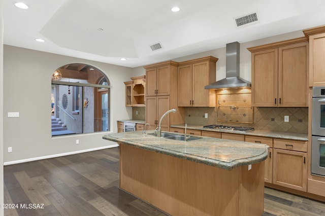 kitchen with sink, an island with sink, wall chimney exhaust hood, stainless steel appliances, and dark hardwood / wood-style flooring