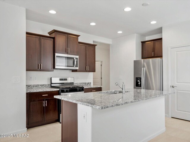 kitchen with a kitchen island with sink, dark brown cabinets, a sink, light stone counters, and stainless steel appliances