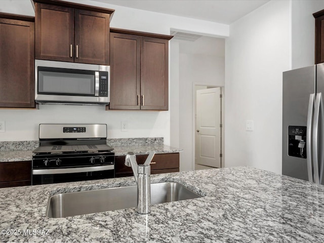 kitchen featuring stainless steel appliances, dark brown cabinetry, light stone counters, and a sink