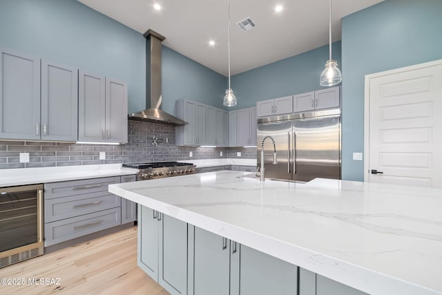 kitchen featuring stainless steel appliances, light stone countertops, wall chimney range hood, beverage cooler, and gray cabinets