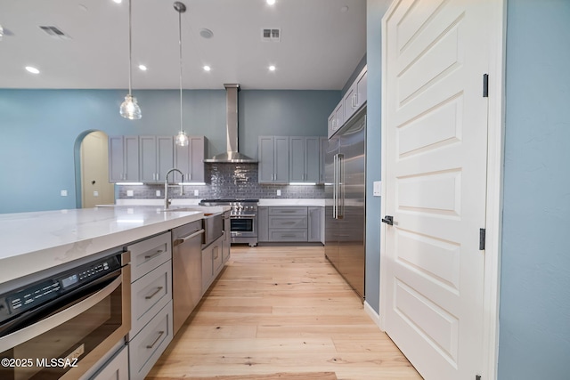 kitchen featuring high end appliances, light stone counters, wall chimney range hood, decorative light fixtures, and gray cabinets