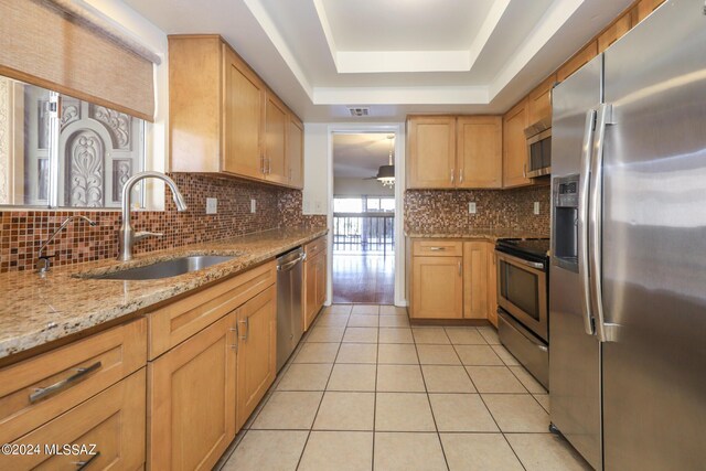 kitchen with appliances with stainless steel finishes, light stone counters, a tray ceiling, sink, and light tile patterned floors