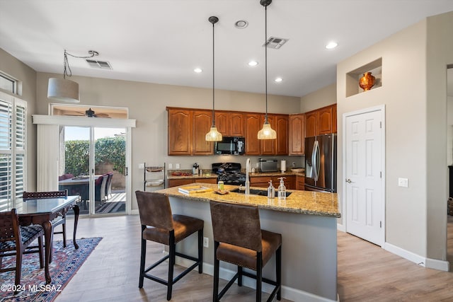 kitchen with pendant lighting, light hardwood / wood-style flooring, black appliances, a center island with sink, and light stone countertops