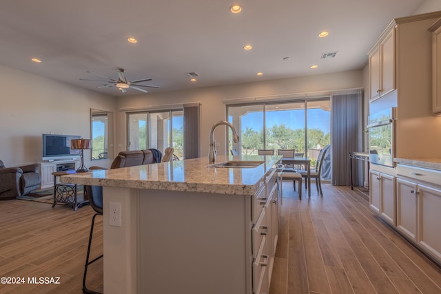 kitchen with light stone counters, ceiling fan, sink, light hardwood / wood-style flooring, and an island with sink