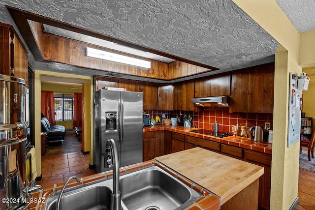 kitchen with a textured ceiling, stainless steel fridge with ice dispenser, tile countertops, range hood, and decorative backsplash