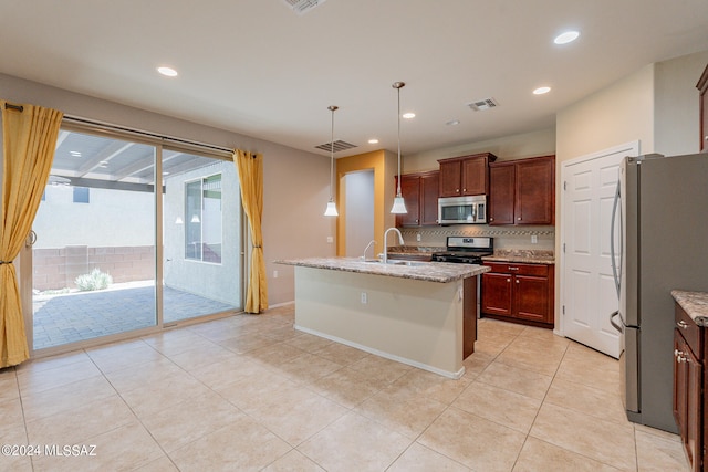 kitchen featuring stainless steel appliances, light stone counters, an island with sink, decorative light fixtures, and decorative backsplash