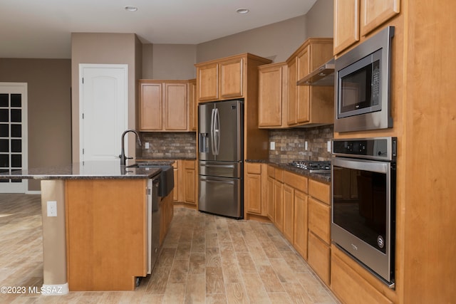 kitchen with appliances with stainless steel finishes, decorative backsplash, a center island with sink, and light wood-type flooring