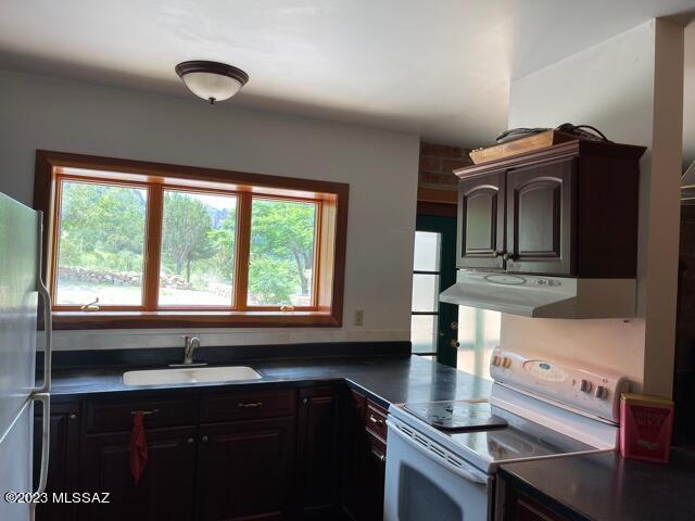 kitchen with white electric stove, under cabinet range hood, a sink, freestanding refrigerator, and dark countertops