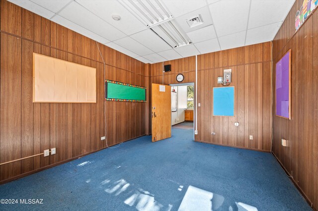 empty room with a drop ceiling, wood walls, and dark carpet