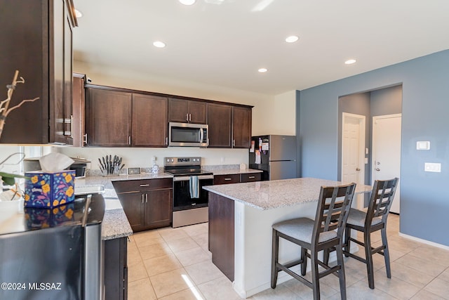 kitchen with light tile patterned floors, appliances with stainless steel finishes, dark brown cabinets, a center island, and light stone countertops