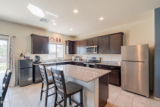 kitchen with dark brown cabinets, stainless steel appliances, a center island, light stone counters, and light tile patterned flooring