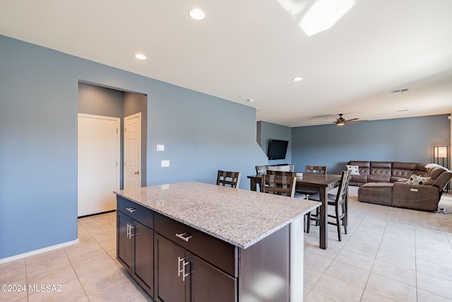 kitchen with a center island, dark brown cabinets, light tile patterned floors, ceiling fan, and light stone countertops