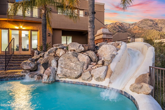 pool at dusk featuring french doors, a mountain view, and pool water feature