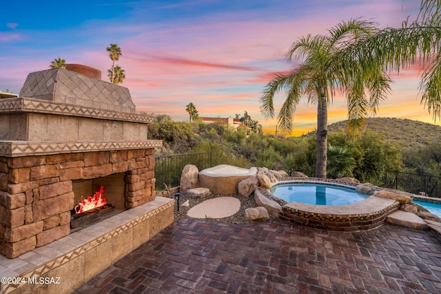 pool at dusk featuring an outdoor stone fireplace and a patio area