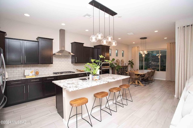 kitchen featuring an island with sink, stainless steel appliances, wall chimney range hood, light stone countertops, and decorative backsplash