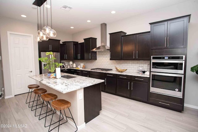 kitchen featuring an island with sink, hanging light fixtures, sink, wall chimney exhaust hood, and appliances with stainless steel finishes