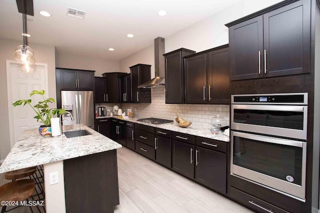 kitchen featuring light stone counters, tasteful backsplash, wall chimney range hood, stainless steel appliances, and a center island with sink