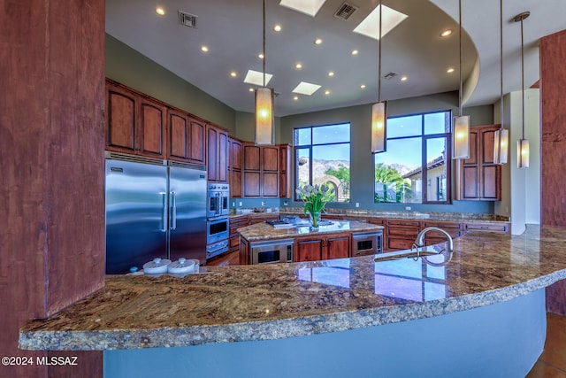 kitchen featuring a skylight, kitchen peninsula, hanging light fixtures, dark stone counters, and built in appliances