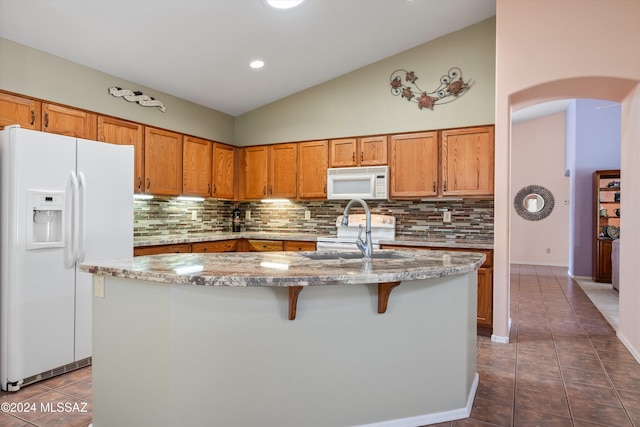 kitchen with dark tile patterned floors, vaulted ceiling, white appliances, a center island with sink, and light stone countertops