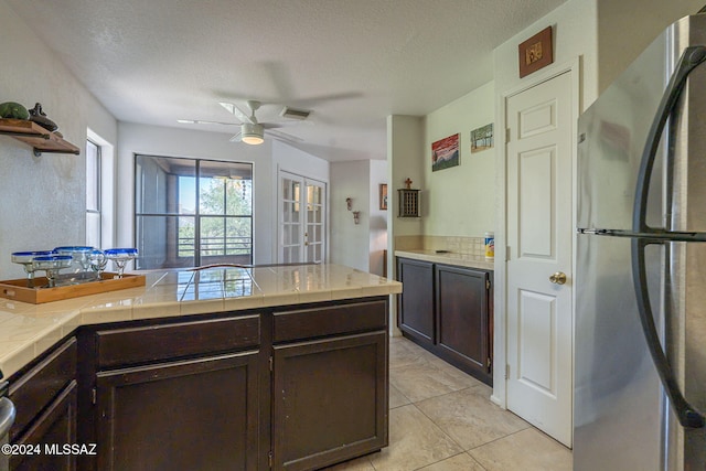 kitchen with stainless steel refrigerator, dark brown cabinets, tile countertops, light tile patterned floors, and a textured ceiling