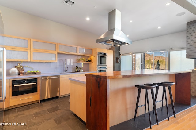 kitchen with tasteful backsplash, island exhaust hood, appliances with stainless steel finishes, a center island, and a kitchen bar