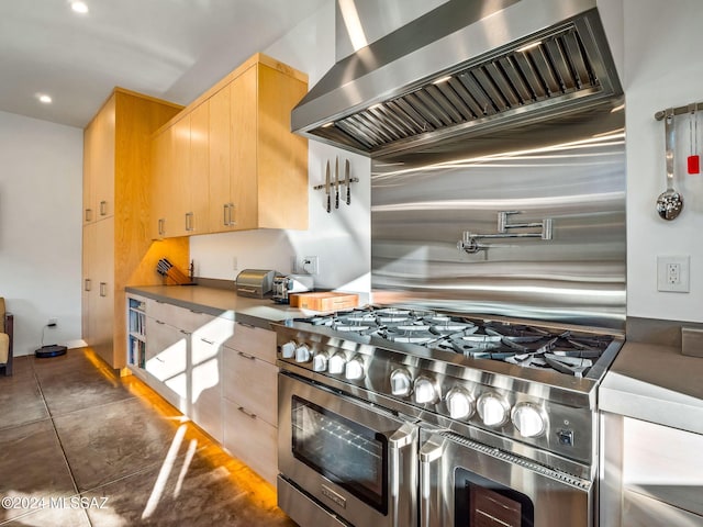 kitchen with dark tile patterned floors, light brown cabinets, range with two ovens, and wall chimney range hood