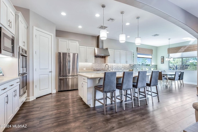 kitchen featuring pendant lighting, wall chimney range hood, an island with sink, white cabinetry, and stainless steel appliances