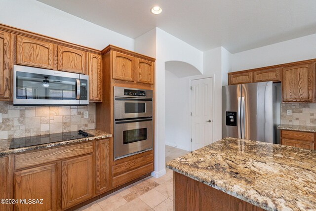 kitchen featuring arched walkways, backsplash, brown cabinets, light stone countertops, and appliances with stainless steel finishes