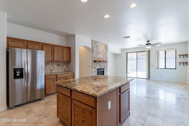 kitchen with ceiling fan, a stone fireplace, backsplash, stainless steel refrigerator with ice dispenser, and a center island