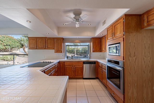 kitchen with ceiling fan, stainless steel appliances, a raised ceiling, tile countertops, and kitchen peninsula