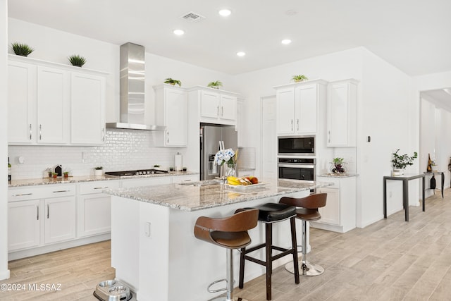 kitchen with wall chimney range hood, light hardwood / wood-style flooring, a center island with sink, white cabinets, and appliances with stainless steel finishes