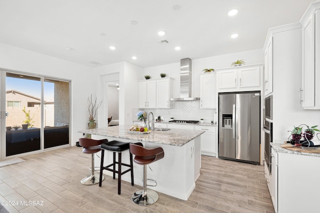 kitchen with wall chimney range hood, light hardwood / wood-style floors, a center island with sink, light stone counters, and appliances with stainless steel finishes