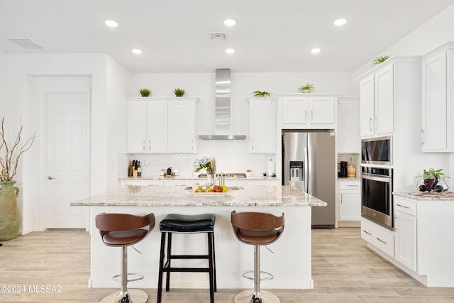 kitchen featuring stainless steel appliances, wall chimney range hood, light hardwood / wood-style floors, and a kitchen island with sink