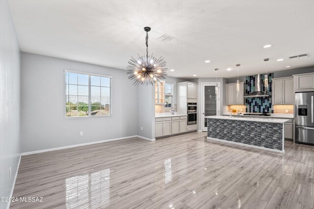 kitchen with pendant lighting, an island with sink, tasteful backsplash, wall chimney exhaust hood, and appliances with stainless steel finishes