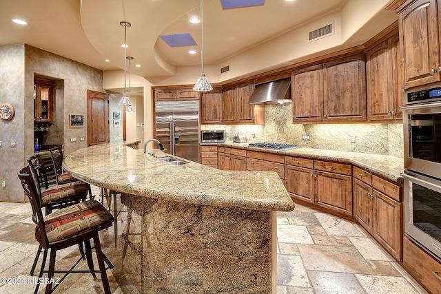 kitchen with decorative backsplash, appliances with stainless steel finishes, wall chimney exhaust hood, a large island with sink, and decorative light fixtures