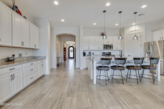 kitchen with white cabinetry, appliances with stainless steel finishes, light stone countertops, hanging light fixtures, and an island with sink