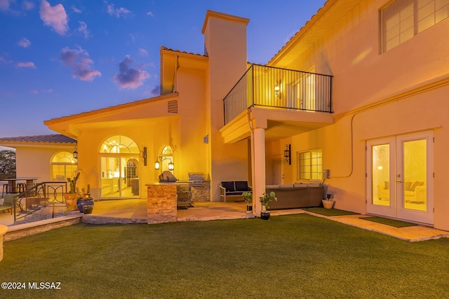 back house at dusk with french doors, a yard, an outdoor kitchen, a balcony, and a patio area