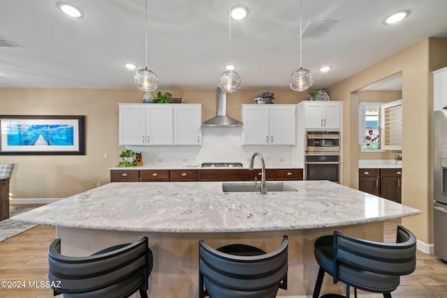 kitchen featuring white cabinetry, pendant lighting, light stone countertops, a kitchen island with sink, and wall chimney range hood