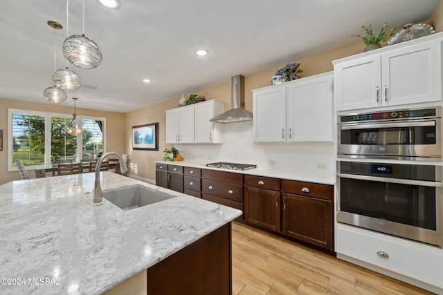 kitchen with wall chimney exhaust hood, sink, hanging light fixtures, appliances with stainless steel finishes, and white cabinets