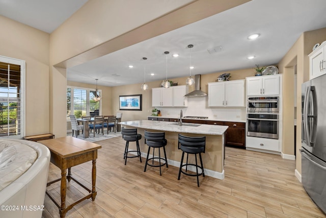 kitchen with white cabinetry, hanging light fixtures, stainless steel appliances, a kitchen island with sink, and wall chimney range hood