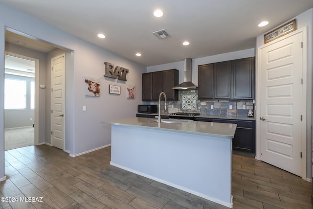 kitchen featuring dark brown cabinetry, wall chimney exhaust hood, tasteful backsplash, an island with sink, and dark hardwood / wood-style floors