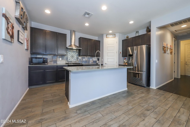 kitchen with appliances with stainless steel finishes, wall chimney exhaust hood, light hardwood / wood-style floors, and a kitchen island with sink