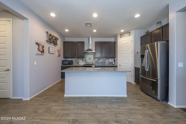 kitchen with an island with sink, stainless steel fridge with ice dispenser, dark brown cabinetry, and wall chimney range hood