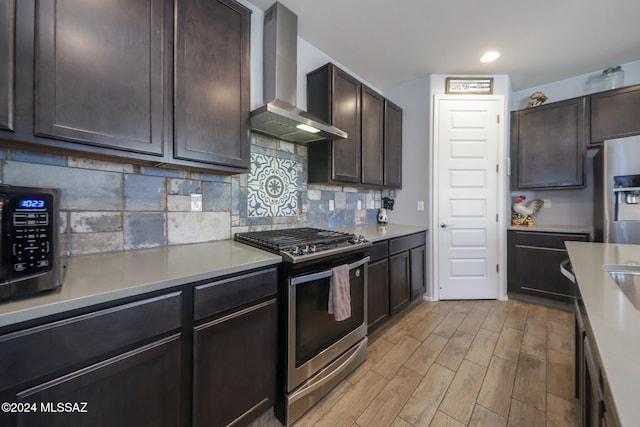 kitchen featuring decorative backsplash, wall chimney exhaust hood, dark brown cabinetry, light wood-type flooring, and appliances with stainless steel finishes