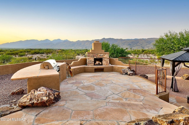 patio terrace at dusk featuring a gazebo, a mountain view, an outdoor stone fireplace, and exterior kitchen