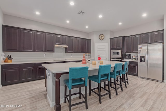 kitchen with appliances with stainless steel finishes, a kitchen breakfast bar, a kitchen island with sink, light hardwood / wood-style flooring, and dark brown cabinetry