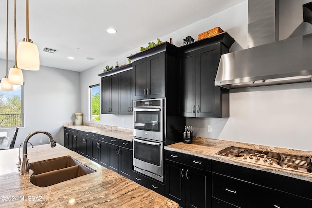 kitchen featuring wall chimney range hood, light stone counters, stainless steel appliances, sink, and decorative light fixtures